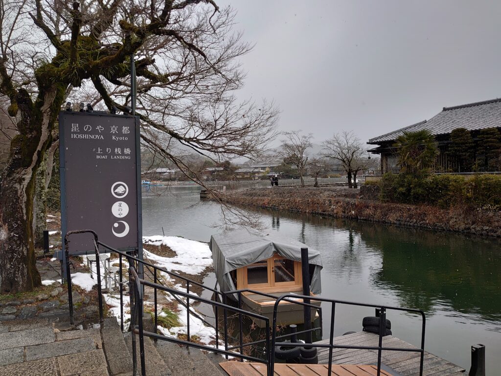 星のや京都の舟乗り場「上り桟橋」。雪景色の中、川沿いに停泊する舟と風情ある案内看板が見える。
