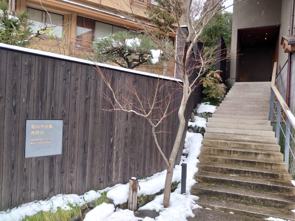 星のや京都の舟待合所の入口。雪の残る石段と木の壁に設置された『舟待合』の案内板が見える。
