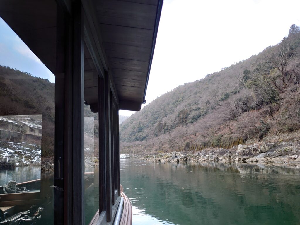 屋形舟から見た保津川の風景。静かな川面と山々に囲まれた穏やかな景色が広がる。舟の窓に景色が映り込んで美しい。