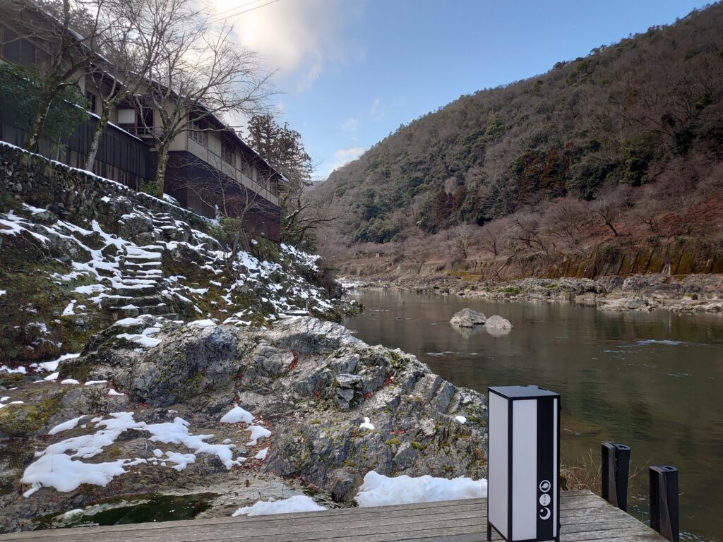 星のや京都の舟着き場から見た宿の外観。雪が残る岩場と、川の流れ、山の緑が調和する自然豊かな風景。