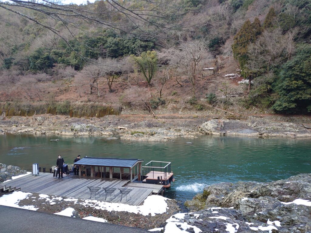 星のや京都の舟着き場の全景。木製の桟橋と屋形舟が停泊しており、周囲の山々と川が冬の静寂を感じさせる。