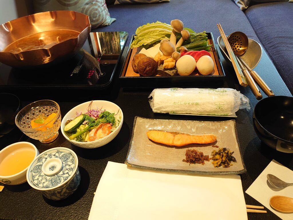 朝鍋の全体図。中央には鮭の焼き物と高菜、左側にサラダと果物、右上には鍋とたっぷりの野菜盛りが配置されている。