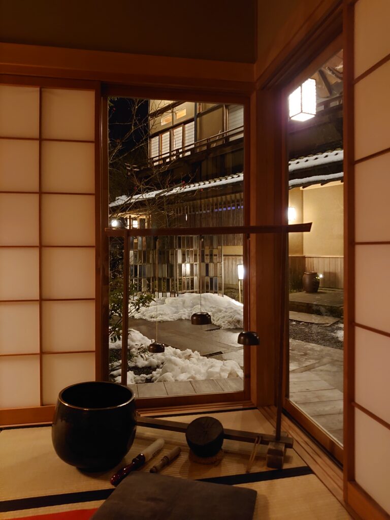 障子越しに雪の中庭を望む和室。畳の上には、仏具をアレンジした音色が印象的な楽器が置かれ、到着時に響いた幻想的な音の余韻を感じさせる空間。