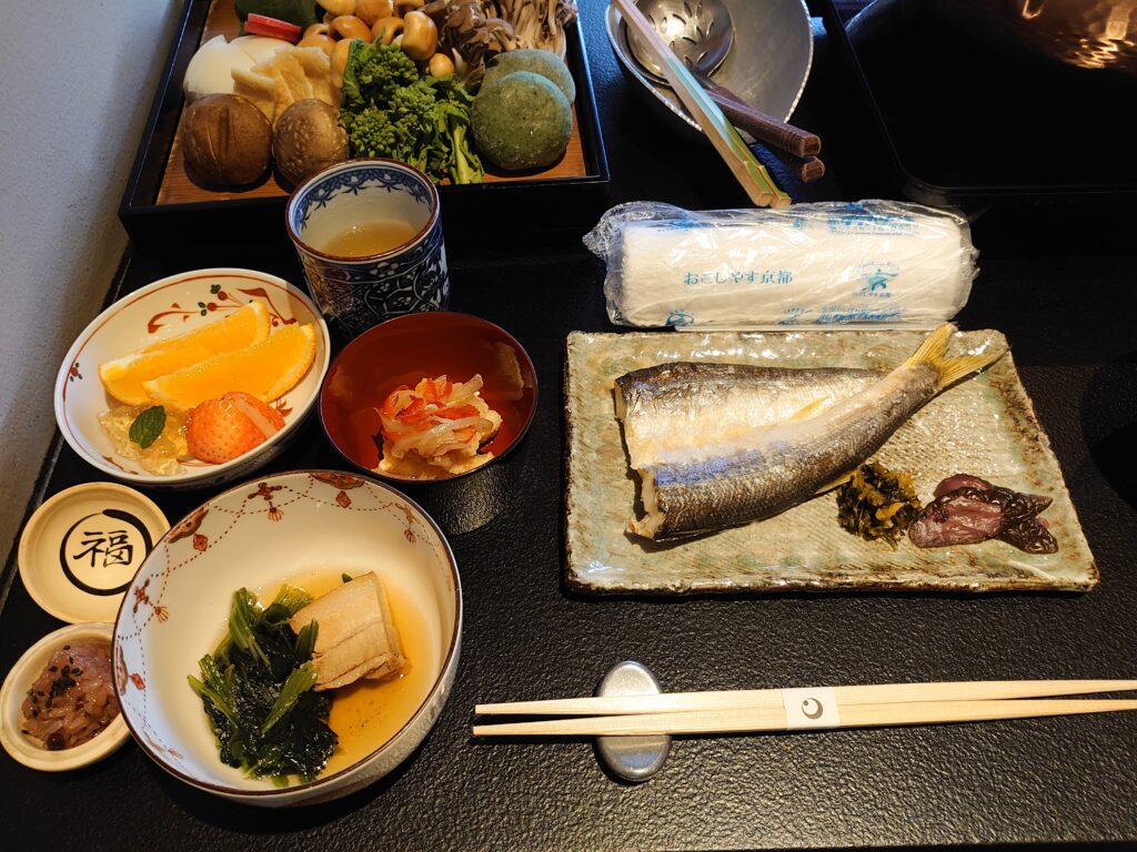 2日目の朝食セット。焼き魚や小鉢料理、フルーツが彩りよく並べられている。鍋の具材は1日目とは違う野菜が用意されている。新婚旅行のお祝い(サービス)のお赤飯もある。